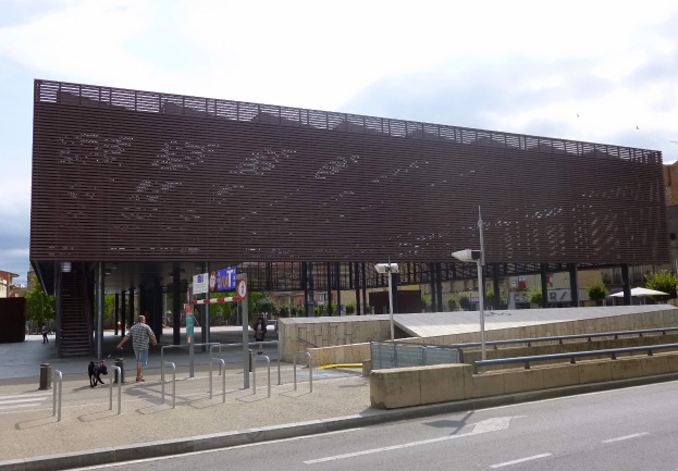 Großes modernes Gebäude mit zahlreichen Fenstern, das als neuer Eingang zur Stadt dient, flankiert von Straßeninfrastruktur und Fußgängern.