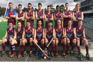 Eine Gruppe von Männern, die auf einem Feld mit Hockey-Schlägern feiern, mit einem Stadion und Zuschauern im Hintergrund unter einem klaren blauen Himmel.