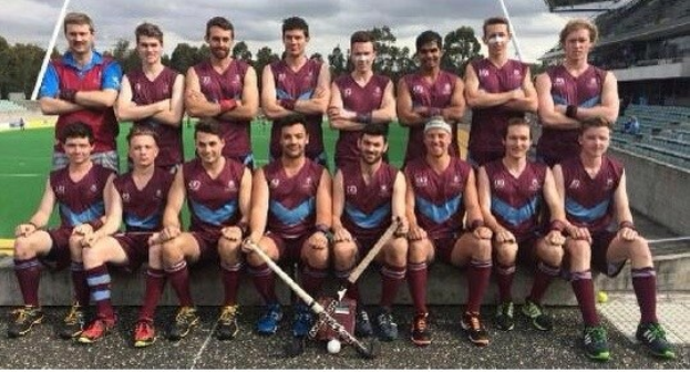 Eine Gruppe von Männern, die auf einem Feld mit Hockey-Schlägern feiern, mit einem Stadion und Zuschauern im Hintergrund unter einem klaren blauen Himmel.