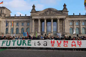 Eine Gruppe von Menschen hält ein Transparent mit der Aufschrift "Zukunft ist ein Menschenrecht" vor dem Reichstag in Berlin, Deutschland, der mit Fenstern, Säulen, Bögen und Flaggenmasten geschmückt ist.