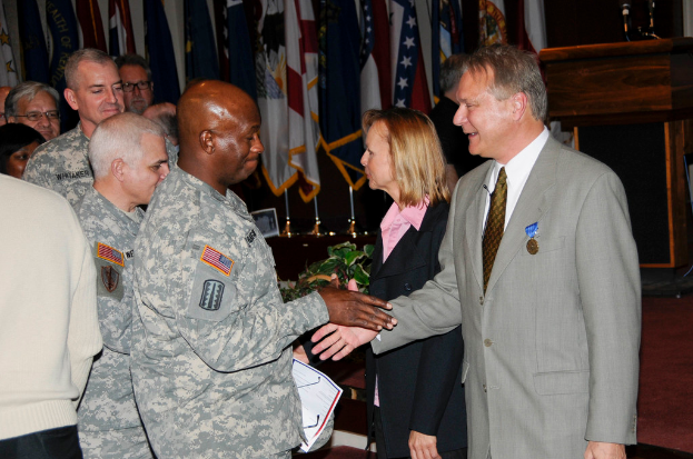 Eine Gruppe von Menschen, die stehen, mit zwei Personen, die sich die Hände schütteln, im Vordergrund, Fahnen im Hintergrund, ein Podium mit einem Mikrofon und eine Topfpflanze in der Nähe.