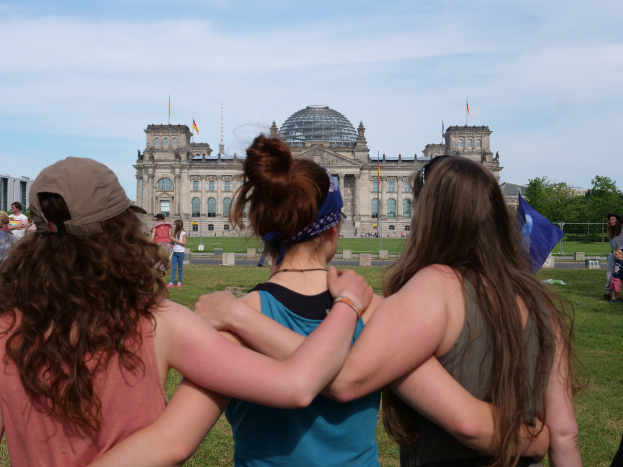Drei Frauen in verschiedenen farbigen Kleidern, eine trägt eine Mütze, stehen vor dem Reichstaggebäude in Berlin, Deutschland, mit Menschen, Fahnen, Bäumen, Wolken und Himmel im Hintergrund.