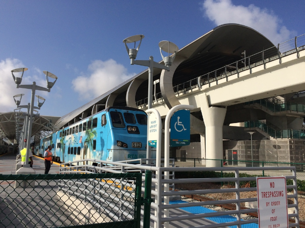 Einige Personen auf einem Bahnsteig mit einem Zug auf der rechten Seite, eine Brücke, ein Zaun und mehrere Pfosten.