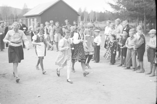 Schwarzes Foto von Kindern, die vor einer Menge mit Fahnen Fußball spielen, mit Bäumen und einem Haus im Hintergrund.