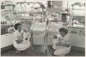 Ein Schwarz-Weiß-Foto von zwei Frauen in einem Krankenhauszimmer, jede hält ein Baby in den Händen, mit Brutkasten und medizinischer Ausrüstung im Hintergrund.
