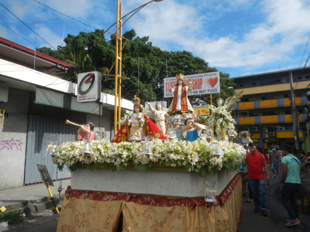 Ein Paradewagen mit Statuen von Menschen, Blumen und anderen Gegenständen, begleitet von Texttafeln, Laternenmasten, Drähten, Gebäuden, Bäumen und einem bewölkten Himmel im Hintergrund.