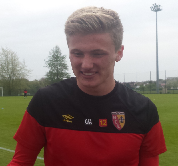 Ein junger Mann steht auf einem Fußballfeld in einem roten und schwarzen T-Shirt, lächelt und hat im Hintergrund ein Tor, eine Umzäunung, Bäume, einen Lichtmast und einen klaren blauen Himmel.