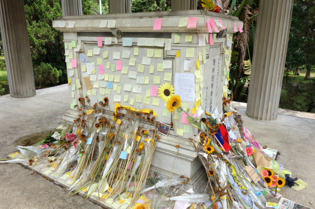 Ein Gedenkstein umgeben von Bäumen und Pflanzen, geschmückt mit Post-it-Notizen und Blumen.