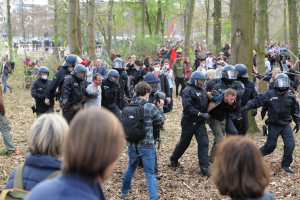 Eine Gruppe von Menschen in Einsatzausrüstung steht vor einer Menge von Menschen mit Helmen und Taschen, mit Bäumen, einer Fahne und Gebäuden im Hintergrund und Blättern auf dem Boden.