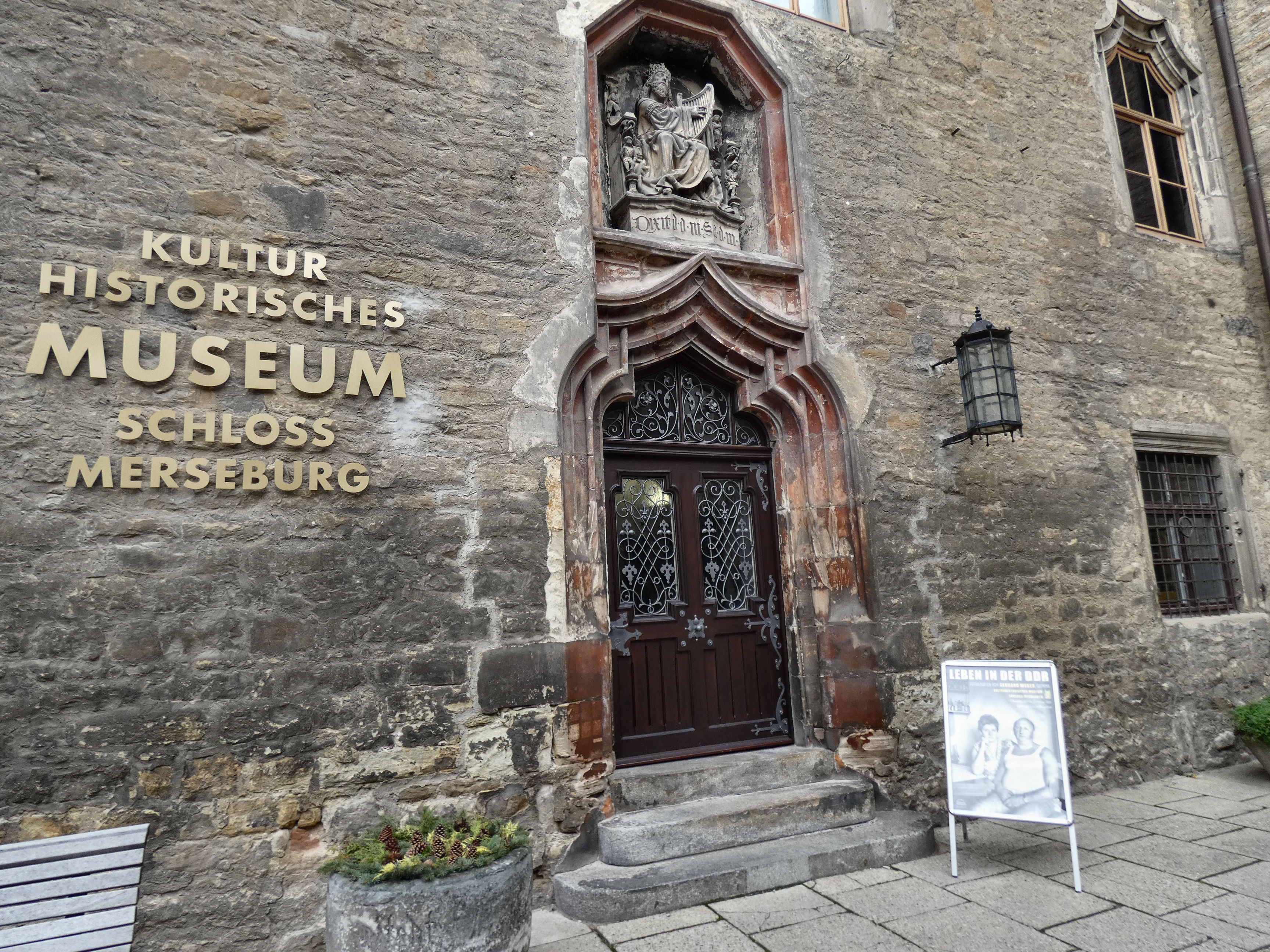 Das Kulturhistorische Museum in Schloss Merseburg, Deutschland, ein Gebäude mit Fenstern, einer Tür, einem Schild, einer Bank, Topfpflanzen und einer Pfeife, unter einem bewölkten Himmel.