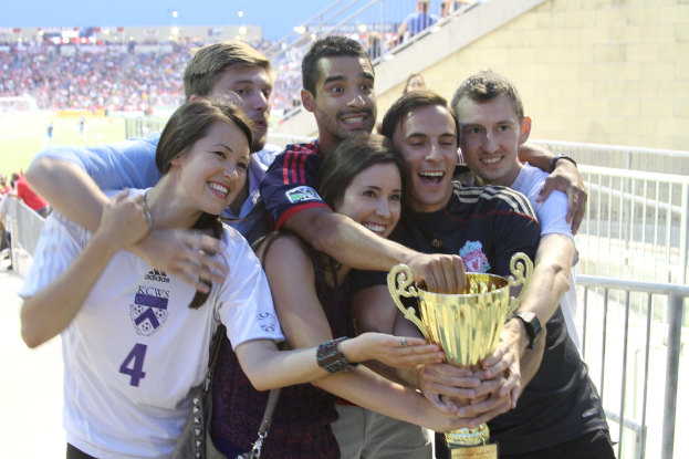 Sechs Personen, zwei Frauen und vier Männer, die eine Trophäe halten und lächeln, posieren für ein Foto vor einem Eisenzaun, mit einer Wand und einem Zaun oben rechts, Menschen und Lichter im Hintergrund, möglicherweise in einem Stadion.