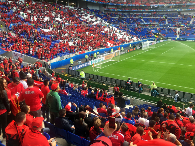 Eine große Menschenmenge in einem hell erleuchteten Stadion schaut einem Fussballspiel zu, mit Zuschauern auf Sitzen und Stehplätzen, Schildern und einem Tor im Hintergrund.