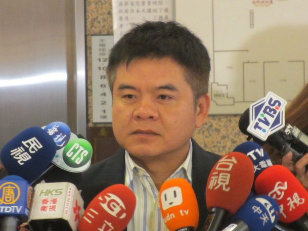 Ein Mann im Anzug und mit Krawatte spricht vor einer Wand mit einer Tafel zu Reportern, während eine Frau rechts eine Kamera hält.