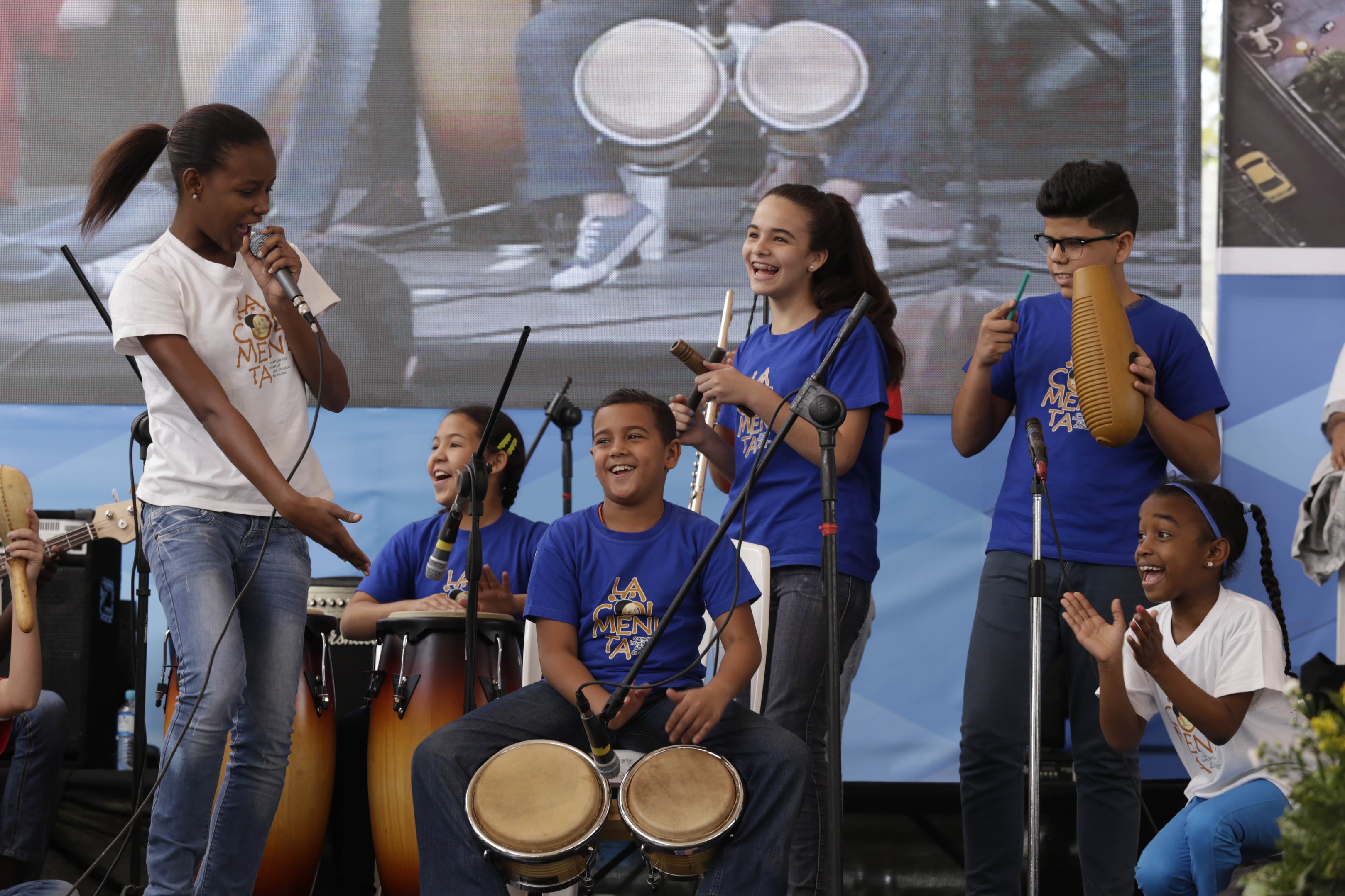 Eine Gruppe von Kindern, die auf einer Bühne Musikinstrumente spielen, mit einer Frau in der Mitte, die ein Mikrofon hält, umgeben von Mikrofonstativen, einem Banner im Hintergrund und Pflanzen auf der rechten Seite.