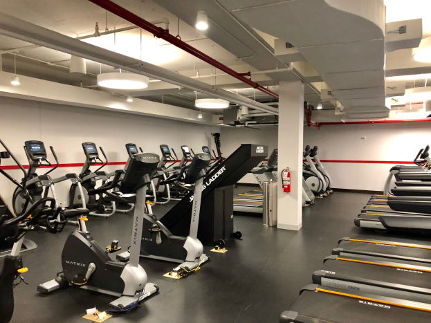 Ein Fitnessstudio-Interieur mit einer Reihe von Laufbändern und Ellipsentrainern, einer Säule, einem Feuerlöscher an der Wand und Deckenleuchten, das sich in einem Fitnessstudio in New York City befindet.