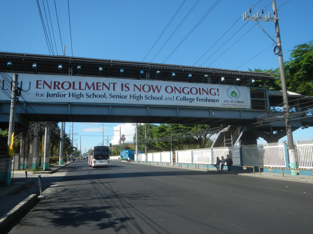 Ein Bus fährt unter einer Brücke mit einem Schild "Jetzt anmelden" vorbei, während Fußgänger darunter gehen und Strommasten die Straße säumen, mit Bäumen und einem klaren Himmel im Hintergrund.