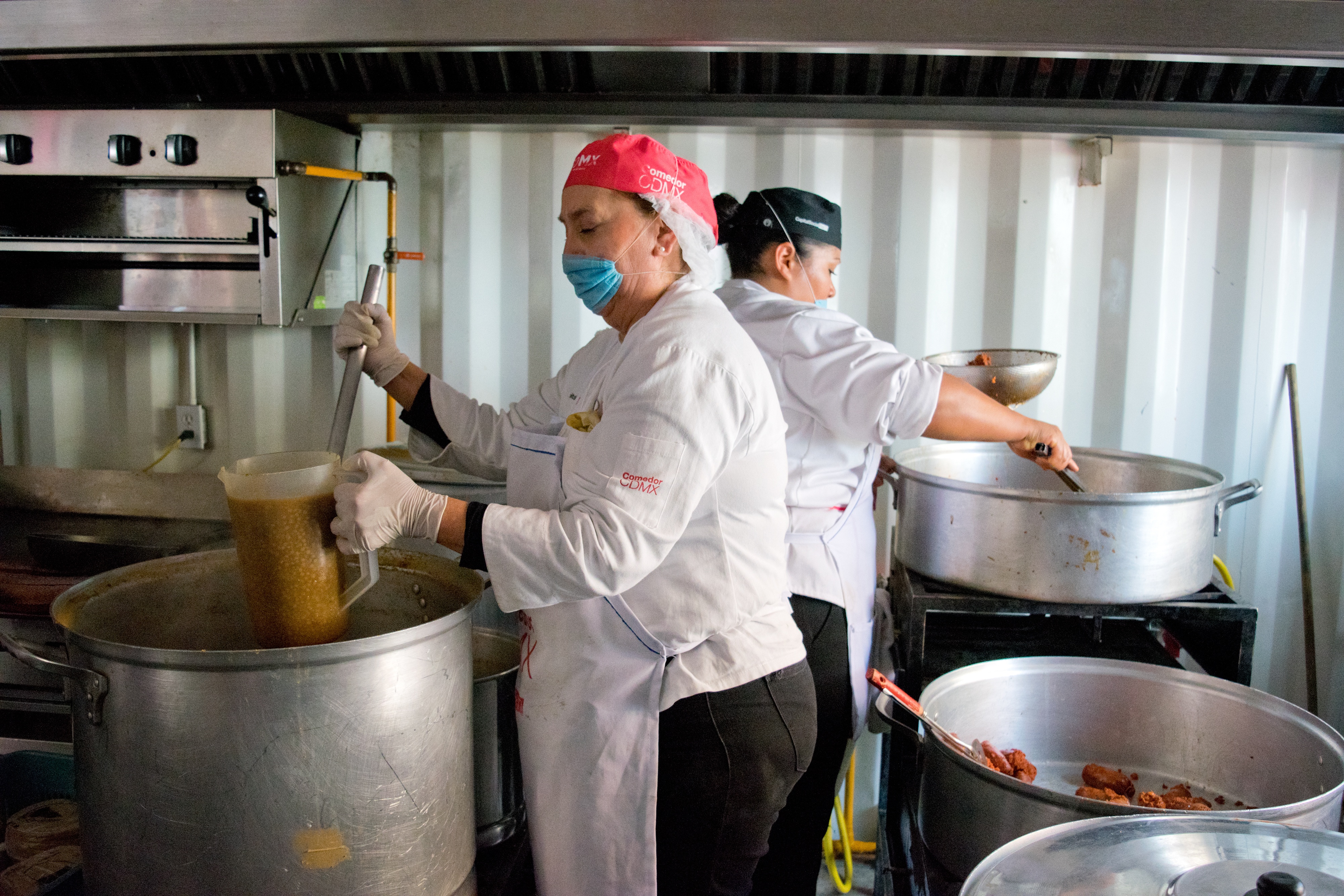 Zwei Frauen in weißen Schürzen, Masken, Kappen und Handschuhen bereiten Essen auf einem Herd mit verschiedenen Utensilien zu, eine hält ein Glas und einen Löffel, vor einem neutralen Hintergrund.