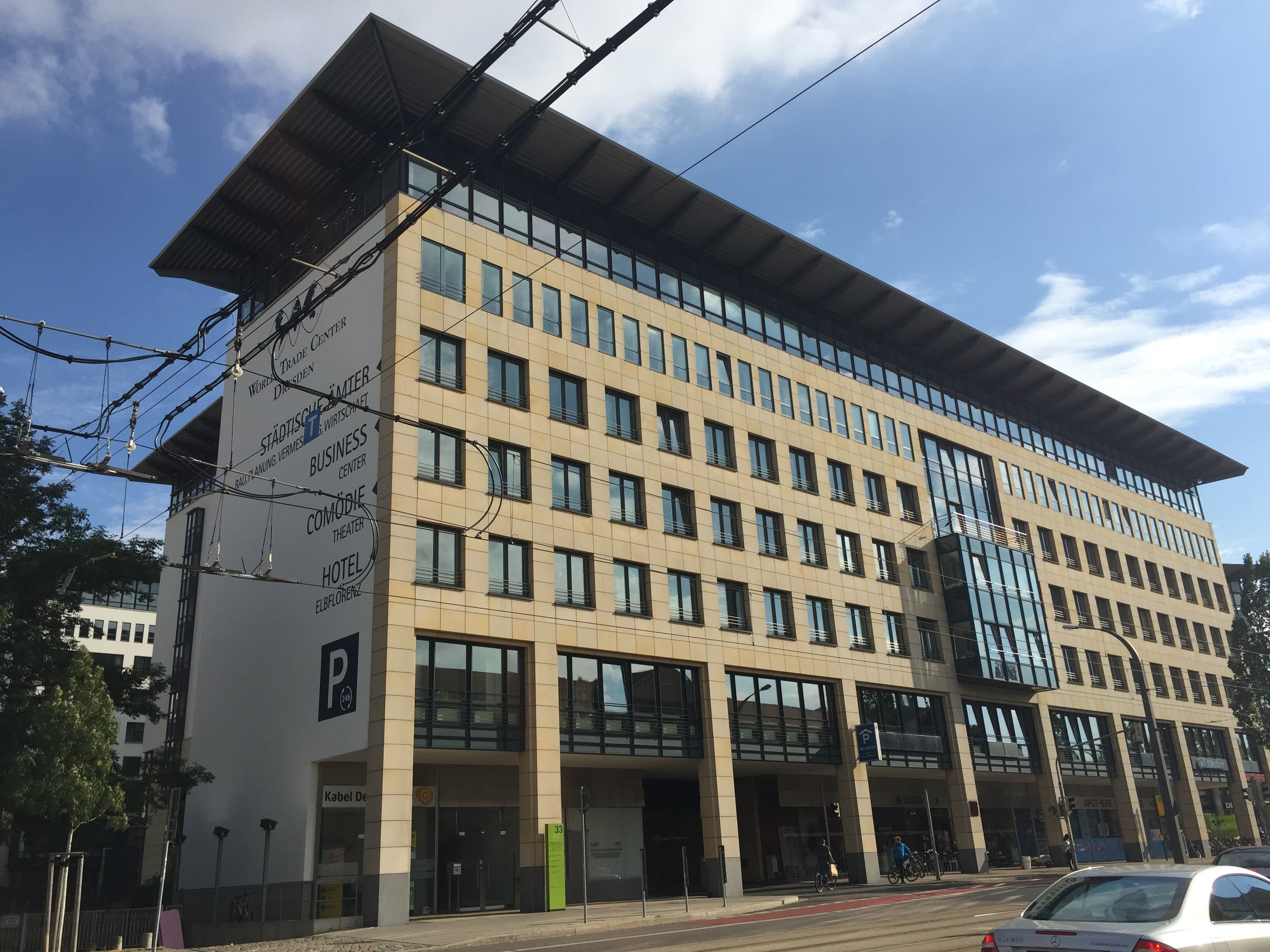 Großes modernes Gebäude mit vielen Fenstern, beschriftet mit "Stadthalle Center for Business", umgeben von Bäumen und städtischer Infrastruktur, mit Fußgängern und Fahrzeugen im Vordergrund unter einem bewölkten Himmel.