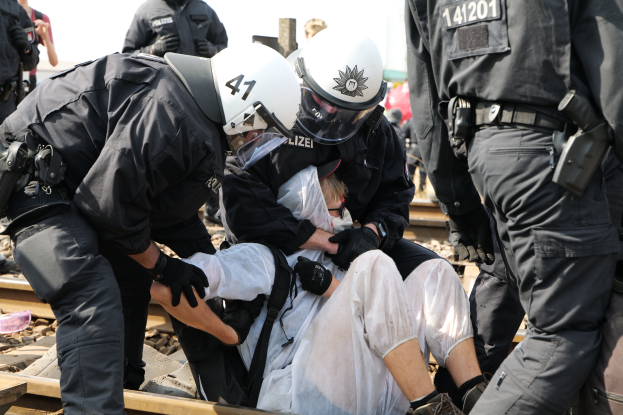 Eine Gruppe von Polizisten in schwarzer Einsatzkleidung verhaftet einen Mann in einem weißen Kleid auf einem Bahnsteig, mit anderen Menschen im Hintergrund.