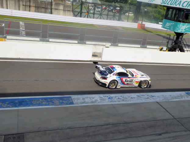 Ein Porsche 911 GT3 RSR fährt auf einer Rennstrecke mit einer Umrandung, Gras, Bäumen, Gebäuden und einem klaren blauen Himmel im Hintergrund, mit einer Texttafel auf der rechten Seite, die die Teilnahme an den 24 Stunden von Le Mans anzeigt.