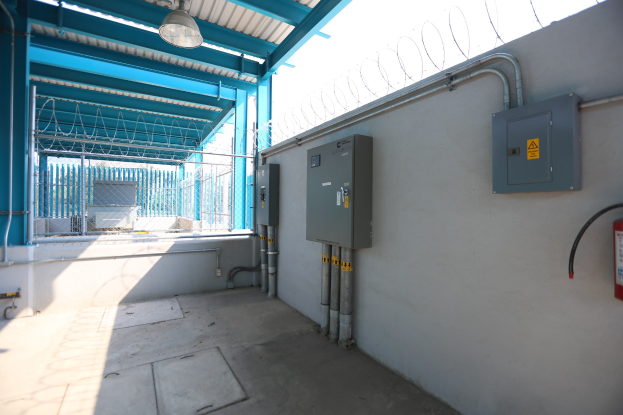 Ein Raum mit einem Feuerlöscher auf der rechten Seite, elektrischen Schaltern und Rohren an der Wand, einer Deckenleuchte und einem Zaun im Hintergrund, was auf eine elektrische Umspannstation hindeutet.
