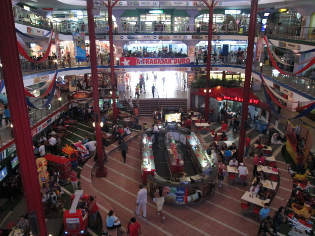 Ein belebtes Einkaufszentrum mit Säulen, Menschen, Tischen, Fliesenboden, Flaggen, Geschäften, einem Banner, Treppen und einem Namensschild.