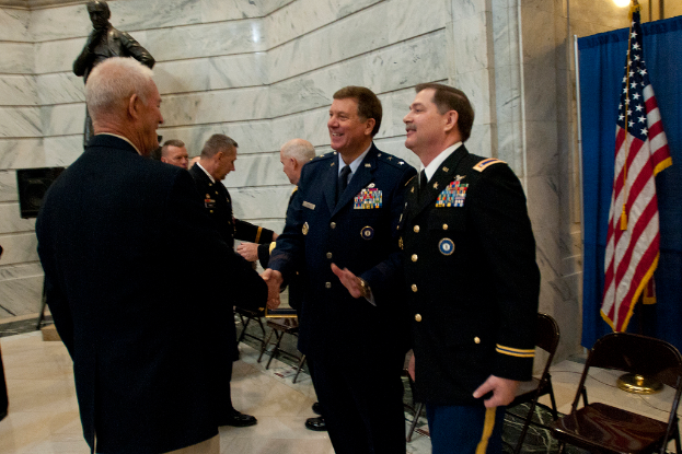 Menschen schütteln Hände mit Stühlen dahinter, Statue neben einer Wand im Hintergrund und Flagge und Vorhang auf der rechten Seite.
