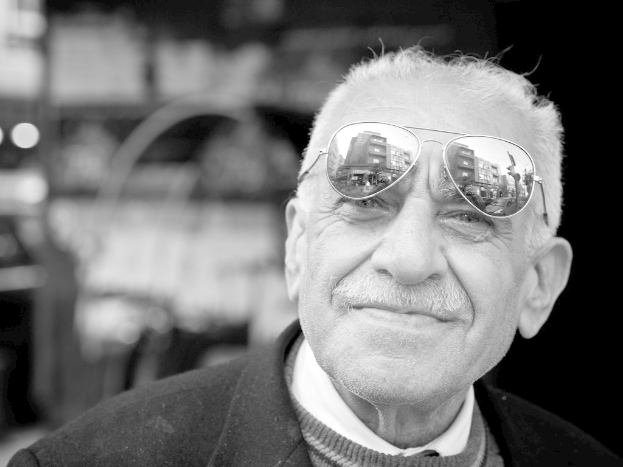 Ein Mann mit einem lachenden Gesicht, der eine Brille trägt, vor einem unscharfen Hintergrund in einem Schwarz-Weiß-Bild.