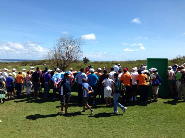 Gruppe von Menschen auf einem grünen Feld bei einem Golfturnier, einige tragen Mützen und tragen Taschen, mit Bäumen, Pflanzen, einem bewölkten Himmel und einem Gewässer im Hintergrund.