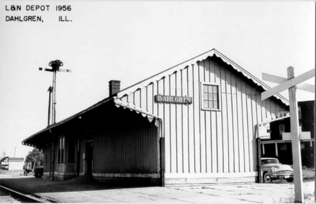 Ein Schwarz-Weiß-Foto eines Bahnhofs in Dahlgren, Illinois aus dem Jahr 1956, das ein Haus mit Fenstern, ein geparktes Fahrzeug, Pfosten, Bäume und einen Himmel im Hintergrund zeigt, mit Text oben und unten im Bild.
