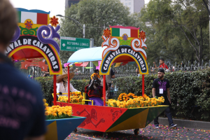 Eine Gruppe von Menschen auf einem blumengeschmückten Festwagen bei einem Umzug, umgeben von einem Metallzaun, Schildern, einer Straßenlaterne, einer Metallabsperrung, Pflanzen, Bäumen und einem Gebäude mit Fenstern unter einem bewölkten Himmel.