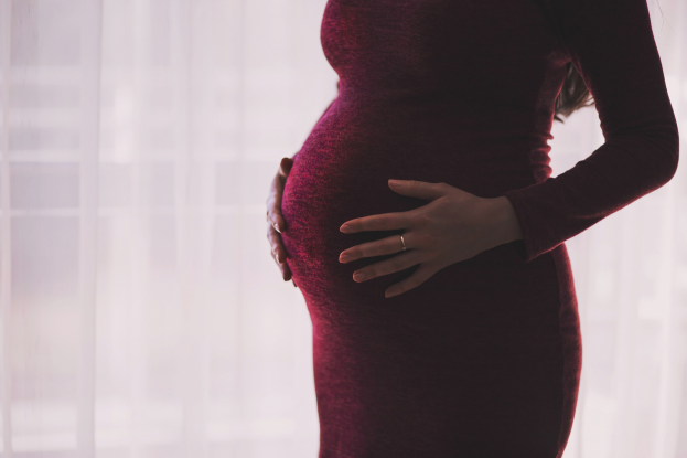 Eine schwangere Frau in einem roten Kleid steht neben einem Fenster, Hände auf ihrem Bauch, mit dem Hintergrund leicht unscharf.
