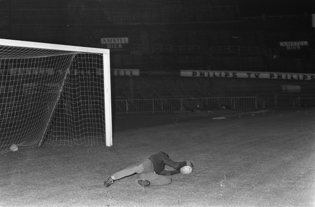 Ein Schwarz-Weiß-Foto eines Fußballspielers, der auf dem Boden liegt und einen Ball vor einem Torpfosten hat, umgeben von einem Zaun und Brettern mit Text im Hintergrund.