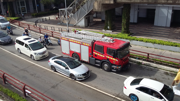 Feuerwehrfahrzeug fährt auf einer Straße mit Motorrädern, Autos und Fußgängern, gesäumt von Geländern und Gebäuden im Hintergrund.