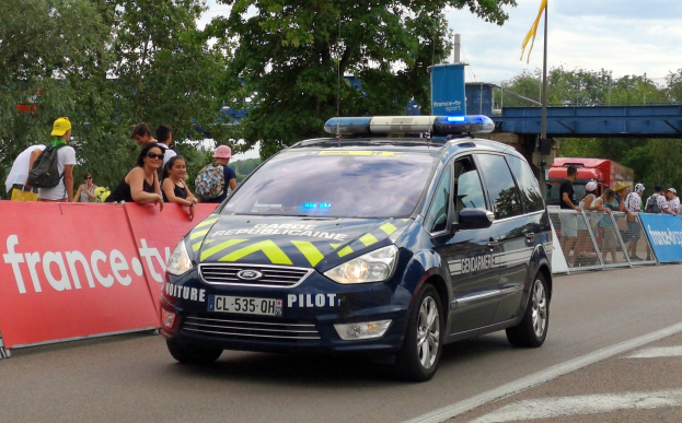 Polizeiauto fährt an einer Menge mit Schildern, Gelöndern, Bäumen, einer Brücke, einer Flagge und einem bewölktem Himmel vorbei.