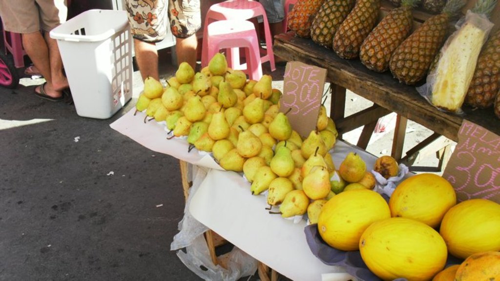 Eine Marktszene mit verschiedenen Fruächten auf einem Gestell, einigen Hockern, ein paar Menschen und zwei Preiszetteln.