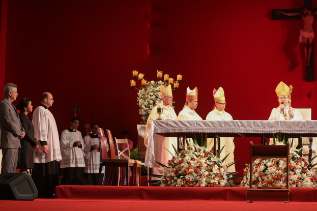 Vier Personen mit Kerzen an Tischen, eine hält ein Mikrofon, Stühle, Lautsprecher, Blumen und heilige Kreuze an der Wand im Hintergrund.
