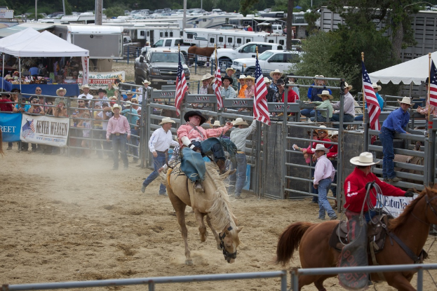 Zwei Menschen auf Pferden bei einem Wettbewerb namens Charreada, umgeben von Gruppen von Menschen, Banner und Fahnen, Pavillonzelten, geparkten Fahrzeugen und Bäumen im Hintergrund.