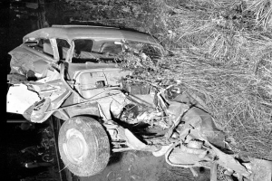 Ein Schwarz-Weiß-Foto eines schwer beschädigten Autos am Straßenrand mit einer eingedrückten Front, zersplitterten Fenstern, einem Baum im Hintergrund und verstreuten Gegenständen um die Unfallstelle.