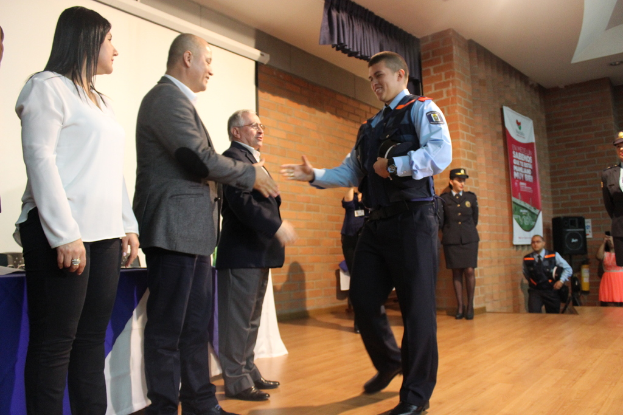 Eine Gruppe von Menschen steht auf einer B√ľhne mit einem Banner, Lautsprecher, Tisch, Bildschirm und Vorh√§ngen im Hintergrund, eine Person in Uniform h√∂lt eine M√ľtze, und eine Frau in einem weißen Hemd.