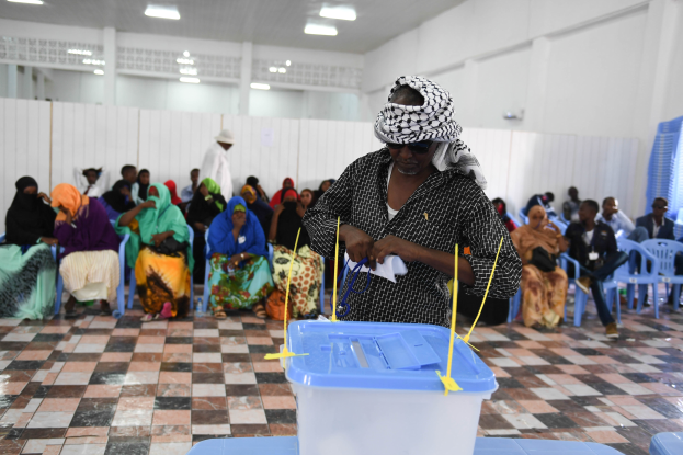 Eine Frau gibt ihre Stimme in einer Wahlurne in Mogadischu, Somalia ab, vor einem Tisch mit einer Box darauf stehend, umgeben von sitzenden Personen und einer Person im Hintergrund, mit Deckenlampen, die den Raum beleuchten.