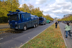 Eine Gruppe von Polizeifahrzeugen, die auf der Seite einer Straße geparkt sind, mit einer Person, die auf der rechten Seite ein Fahrrad fährt, vielen Menschen, die Fahrräder halten, Gras und trockene Blätter auf dem Boden und Bäume, Gebäude und ein bewölkter Himmel im Hintergrund.