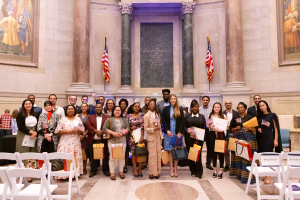 Eine Gruppe von Menschen, wahrscheinlich Absolventen, steht vor einem Gebäude und hält Taschen und Urkunden in den Händen, mit Stühlen auf beiden Seiten, Fahnen, Säulen und Foto Rahmen im Hintergrund.