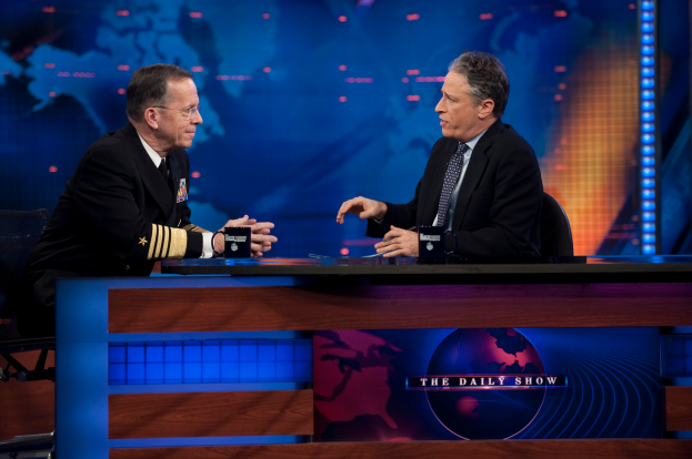 Zwei Männer in schwarzen Blazern, Hemden und Krawatten sitzen an einem Tisch mit Mikrofonen zwischen ihnen, in ein Gespräch vor einem Fernseher und einem blauen Hintergrund vertieft.