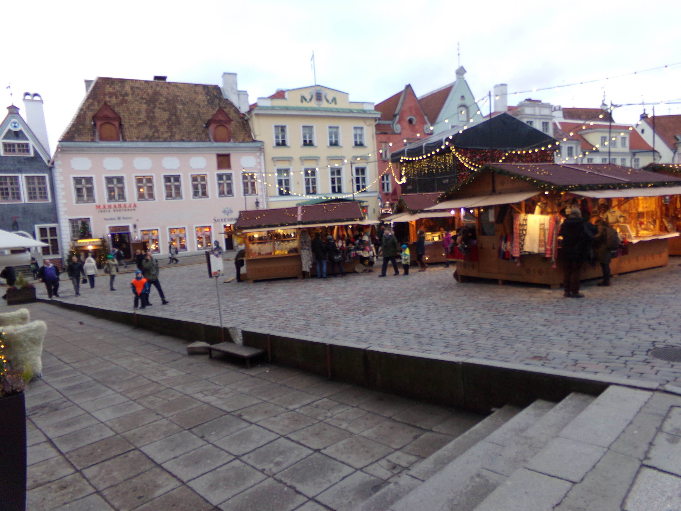 Ein belebter Weihnachtsmarkt in Tallinn, Estland mit Menschen um geschmützte Stände, festliche Lichter, Topfpflanzen, Treppen führend und bewölktem Himmel mit Gebäuden im Hintergrund.