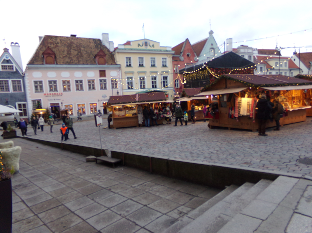 Ein belebter Weihnachtsmarkt in Tallinn, Estland mit Menschen um geschmützte Stände, festliche Lichter, Topfpflanzen, Treppen führend und bewölktem Himmel mit Gebäuden im Hintergrund.
