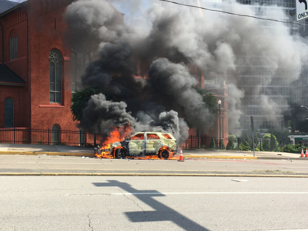 Ein Auto in Flammen an der Straßenseite mit Rauch, der aus ihm aufsteigt, umgeben von Gebäuden, Bäumen und anderen urbanen Elementen unter einem klaren Himmel.