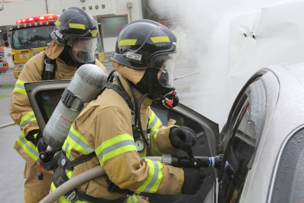 Feuerwehrleute in Schutzausröstung löschen ein brennendes Auto mit einem Schlauch, während Rauch aufsteigt, mit Fahrzeugen und einem Gebäude im Hintergrund.