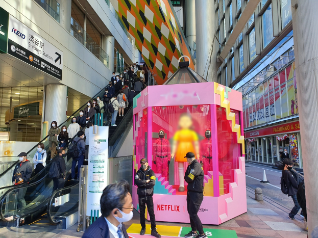 Eine Gruppe von Menschen steht vor einem Gebäude mit Glaswänden und Texttafeln, einige tragen Masken, während andere eine Rolltreppe benutzen; ein pinker Kasten in der Mitte zeigt Schaufensterpuppen und trägt den Text "Netflix's new Netflix booth in Tokyo."