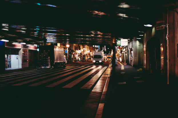 Eine Stadtstraße bei Nacht mit Fahrzeugen auf der Straße, Gebäuden auf beiden Seiten, beleuchteter Bereich und eine Brücke im Hintergrund.